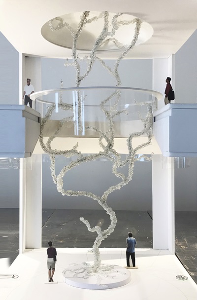 A model of a sculpture designed by artist Maya Lin appears like a branching vertical structure across two stories of an indoor space, populated by model people. The image is paired with a sketch version of the same sculpture.